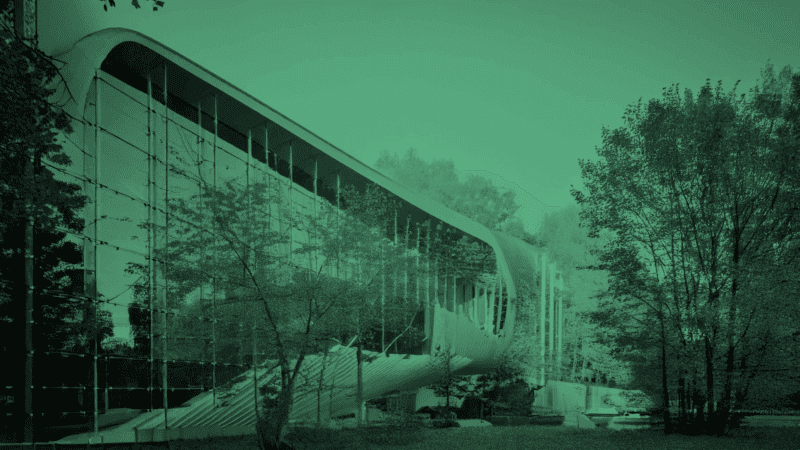 A modern building with a curved glass facade stands in a green-tinted image, surrounded by trees and foliage in a park-like setting.